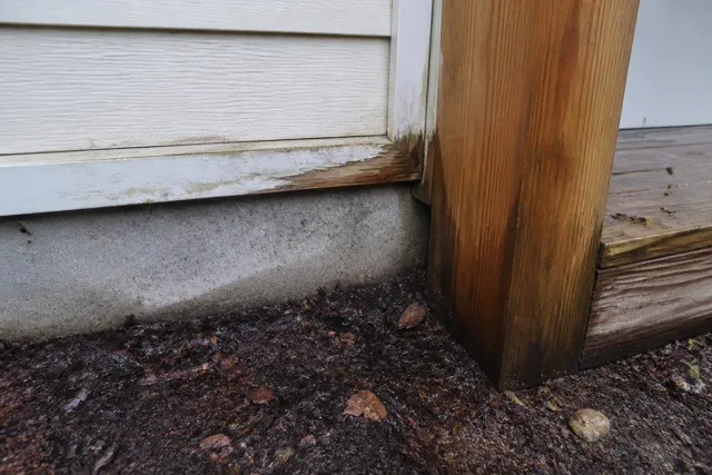 Damp soil and mulch against a house foundation with moisture staining on the wood trim and porch post above
